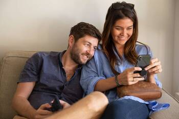 Zwei Personen nutzen Smartphone, junger Mann lehnt sich dabei lächelnd an junge Frau an