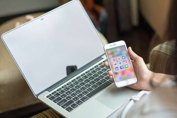 Person mit MacBook auf dem Schoß schaut auf iPhone mit offenem Social Media Ordner