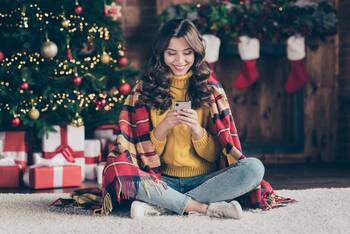 Frau sitzt lächelnd mit einer Decke über den Schultern mit ihrem Smartphone vorm Weihnachtsbaum