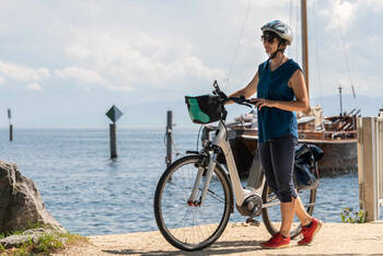 Frau mit Helm steht neben Fahrrad an einem See.