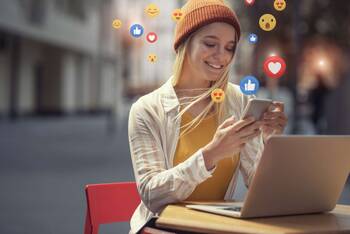 Frau sitzt lächelnd vor Laptop, hält ihr Smartphone in der Hand aus dem Emojis strömen