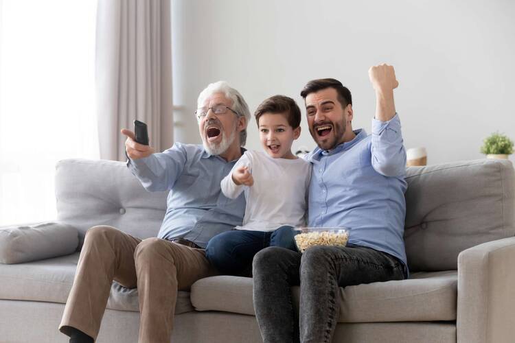 3 Männer aus 3 Generationen sitzen auf Couch und schauen jubelnd Fernsehen