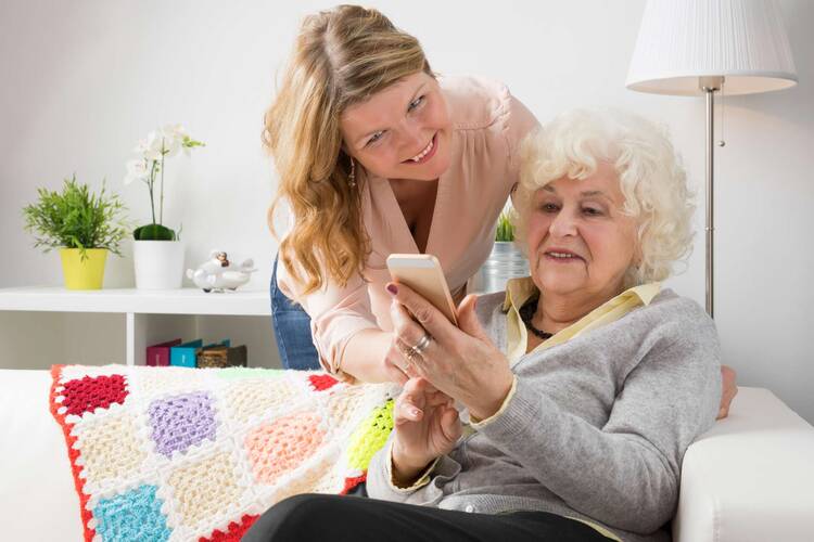 Frau steht hinter älterer, auf Sofa sitzender Frau und erklärt ihr etwas am Handy