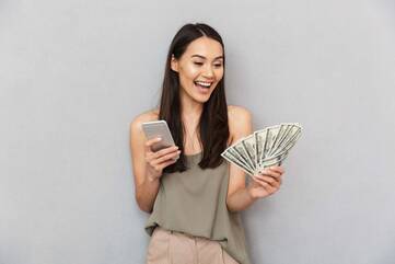 Frau freut sich, hält in einer Hand Geldscheine, in der anderen Smartphone