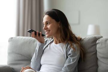 Frau sitzt auf Sofa und spricht in Smartphone-Lautsprecher