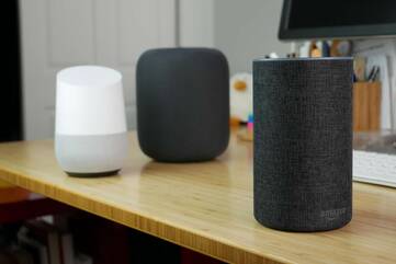 Amazon Echo in der Farbe Anthrazit Stoff steht auf einem Tisch rechts vorne, dahinter (mit Tiefenunschärfe) mittig der Apple HomePod in der Farbe Space Gray und rechts daneben der Google Home in der Farbe Weiß