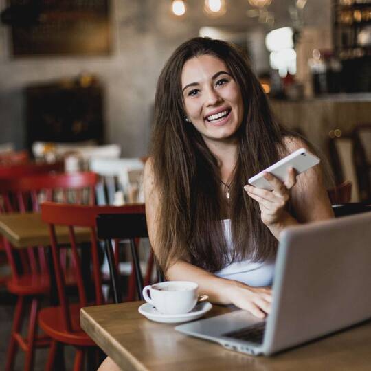 Frau mit Handy in der Hand sitzt lächelnd an Tisch im Café