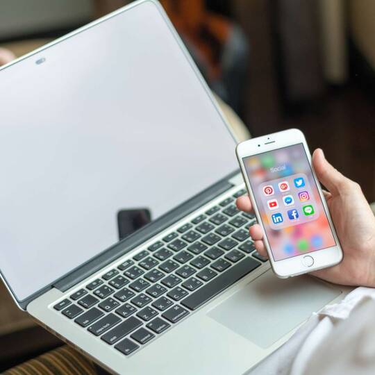 Person mit MacBook auf dem Schoß schaut auf iPhone mit offenem Social Media Ordner