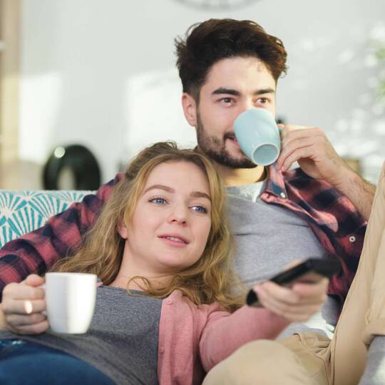 Paar mit Tassen sitzt auf Couch und schaut Fernsehen