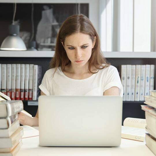 Frau guckt mit offenem Buch in der Hand konzentriert auf ihren Laptop