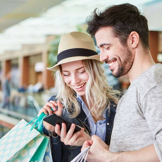Mann und Frau mit Einkaufstaschen gucken gemeinsam lächelnd auf ein Smartphone