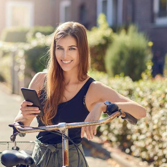 Frau mit Smartphone in der Hand steht lächelnd an Fahrradlenker angelehnt
