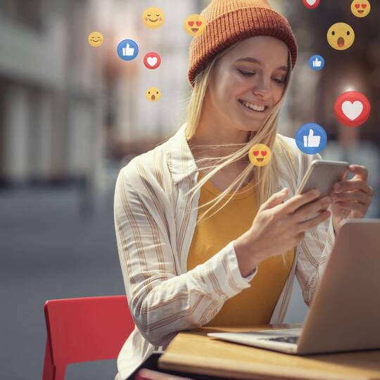 Frau sitzt lächelnd vor Laptop, hält ihr Smartphone in der Hand aus dem Emojis strömen