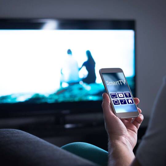 Smartphone wird vor einem Fernseher in der Hand gehalten
