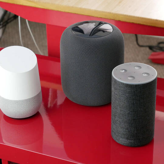 Google Home, Apple HomePod, Amazon Echo stehen nebeneinander auf rotem Tisch