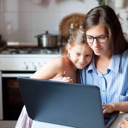 Frau und Kind sitzen in Küche vor Laptop und schauen gemeinsam auf Bildschirm