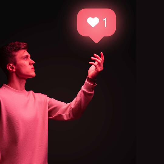 Junger Mann mit ausgestrecktem Arm und rotem "Like"-Zeichen von Instagram vor schwarzem Hintergrund