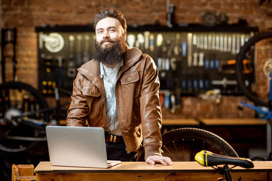 Ein Mann steht in einer Fahrradwerkstatt an einem Tisch und lächelt. Vor ihm steht ein Laptop.