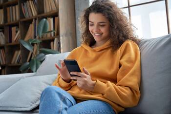 Frau im gelben Pulli auf Sofa tippt auf Smartphone