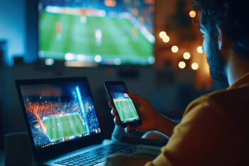 Ein Mann sitzt vor dem Laptop und Fernseher und schaut auf beiden Geräten Fußball.