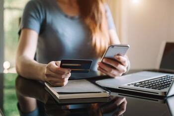 Frau sitzt neben MacBook und Notizblock an Tisch, hält in einer Hand Kreditkarte, in anderer Hand iPhone