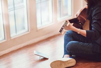 Mensch sitzt im Schneidersitz mit Gitarre auf dem Schoß auf dem Boden mit aufgestelltem Smartphone vor sich