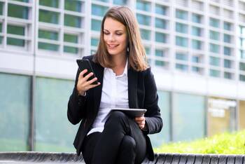 Frau in Business Klamotten sitzt draußen mit Handy in einer Hand und Tablet in anderer Hand