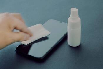 Hand mit Reinigungstuch reinigt Smartphone Display, daneben steht eine kleine, weiße Sprühflasche ohne Etikett