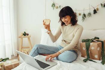 Frau sitzt lächelnd mit Weinglas in der Hand vor Laptop in gemütlicher, weihnachtlicher Umgebung