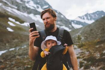Mann mit Kind im Tragegurt in den Bergen schaut auf sein Smartphone