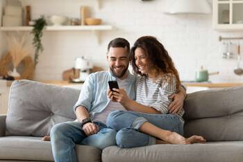 Paar sitzt lächelnd auf Couch und schaut gemeinsam auf ein Smartphone