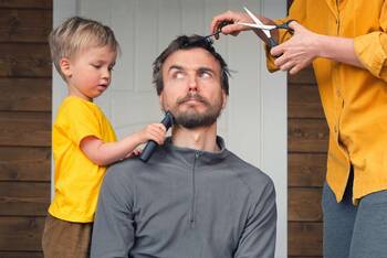 Skeptisch schauender Mann mit Frau und Kind rechts und links, die ihm die Haare schneiden