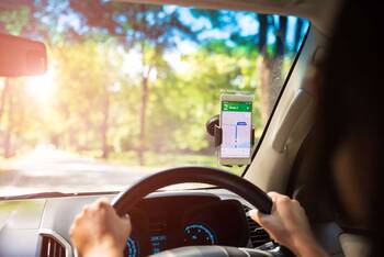 Person lenkt mit beiden Händen Auto, ein Smartphone mit Maps App ist am Halter an der Scheibe