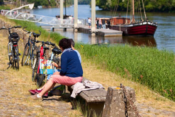 Zwei Personen sitzen auf einer Bank am Rande eines Radweges und schauen in ein Prospekt. Neben Ihnen stehen meherere Fahrräder. Am Radweg führt ein Fluss vorbei. Im Hintergrund ist ein Boot zu sehen.