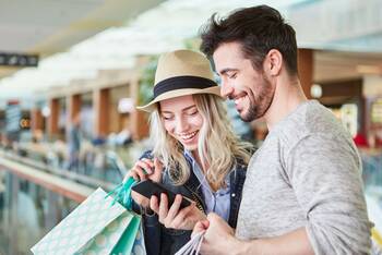 Mann und Frau mit Einkaufstaschen gucken gemeinsam lächelnd auf ein Smartphone