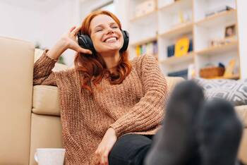 Rothaarige Frau sitzt mit Over Ear Kopfhörern fröhlich Musik hörend auf Sofa