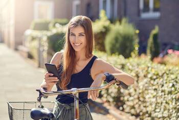 Frau mit Smartphone in der Hand steht lächelnd an Fahrradlenker angelehnt
