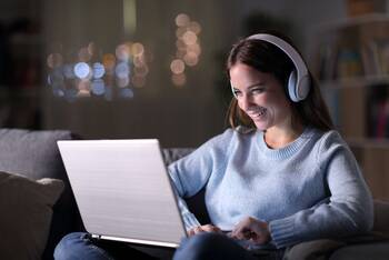 Frau mit großen Kopfhörern sitzt lächelnd vor Laptop in dunkler Umgebung