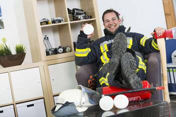 Mann in Feuerwehranzug sitzt lächelnd in Büro, auf Tisch vor ihm liegen Helm und Feuermelder