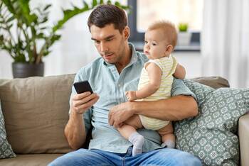Mann mit Baby im Arm sitzt auf Sofa und hält Smartphone in der Hand
