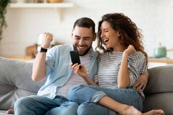 Paar sitzt mit freudigen Gesten auf einem Sofa und guckt lachend auf ein Smartphone