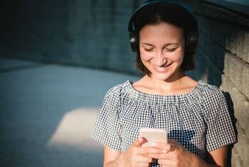 Frau sitzt lächelnd mit Over Ear Kopfhörern vor Handy