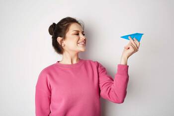 Frau in rosa Pulli hält lächelnd ein blaues Papierflugzeug in der Hand