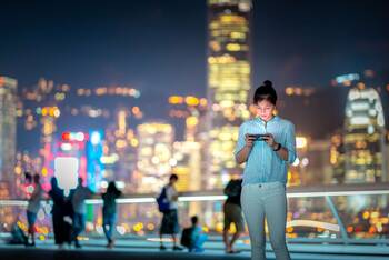 Eine Eine Frau steht vor der Skyline von mehreren Hochhäusern, sie schaut auf ihr Smartphone in ihrer Hand.