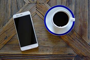 Ein weißes Smartphone auf einem Holztisch. Rechts daneben eine Tasse mit einer schwarzen Flüssigkeit.