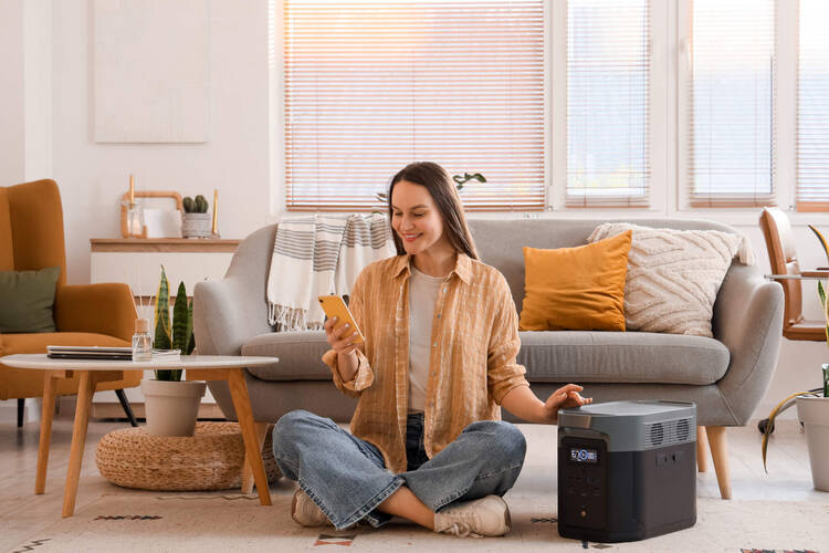 Eine Frau sitzt im Wohnzimmer auf dem Boden und lädt ihr Handy an einer Powerstation auf.