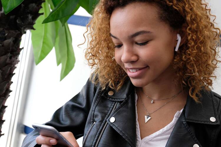 Junge Frau in Lederjacke mit AirPods in den Ohren schaut lächeldn auf Smartphone Display