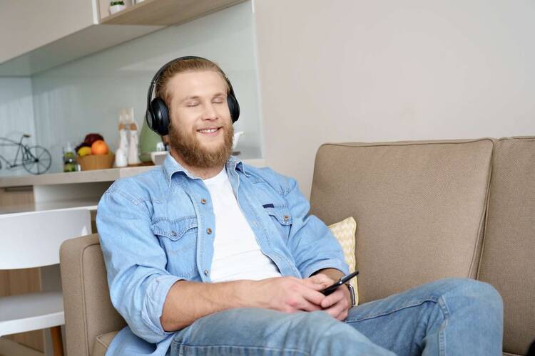 Mann mit Over Ear Kopfhörern sitzt lächelnd mit Smartphone in der Hand auf Sofa