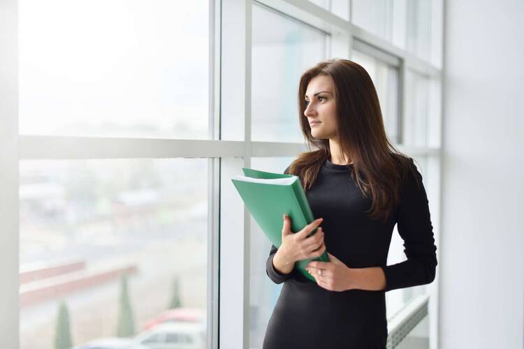 Frau in Business-Klamotten mit Dokumenten in der Hand steht an Fensterfront und schaut nach draußen
