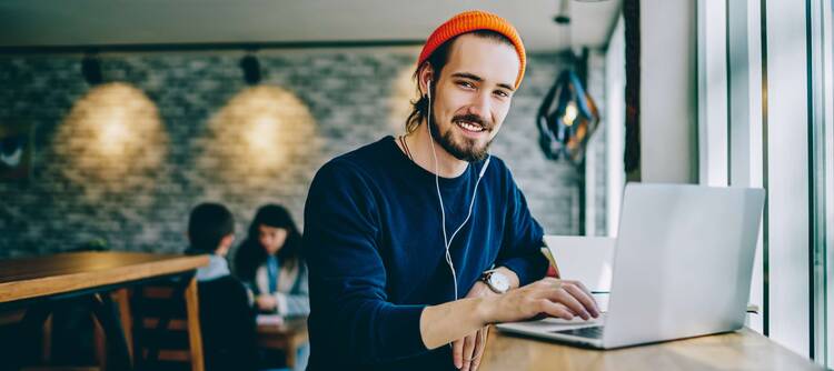 Junger Mann mit Mütze und Kopfhörern sitzt mit Laptop an öffentlichem Ort und lächelt